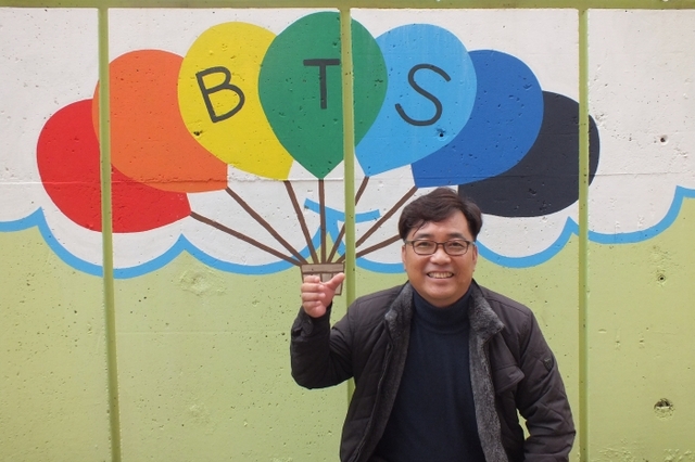 수원조원초등학교 손문성 교감이 학교 담벼락에 직접 그린 방탄소년단을 상징하는 풍선을 배경으로 활짝 웃고 있다.